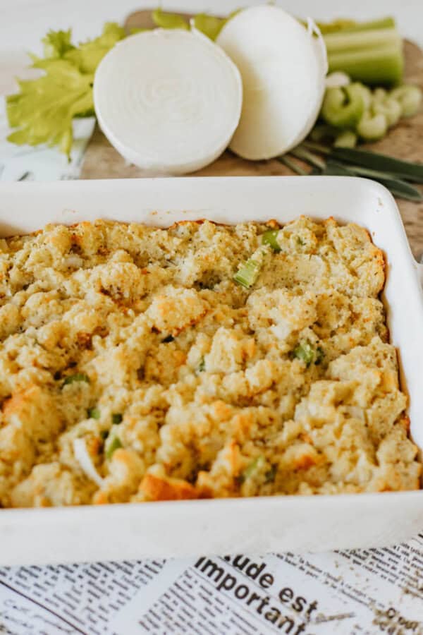 A white casserole dish filled with golden-brown stuffing, garnished with pieces of celery. In the background, theres a halved onion, additional celery stalks, and a newspaper.