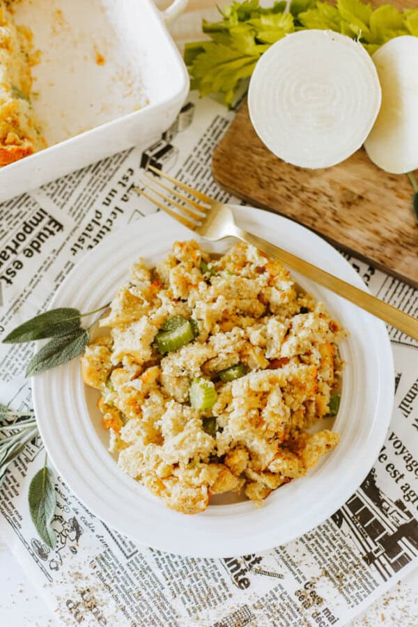 A plate of cornbread stuffing with celery is on a table covered with newspaper-patterned paper. A gold fork rests on the plate. Sage leaves are scattered nearby, alongside sliced onions and fresh celery on a wooden board.