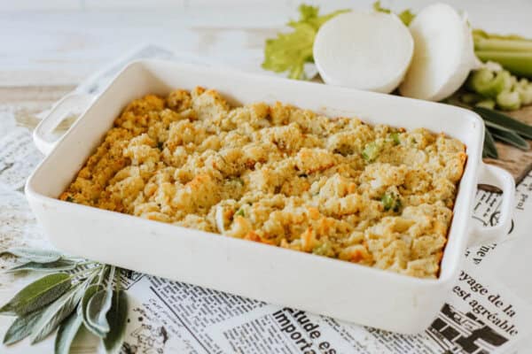 A white casserole dish filled with baked stuffing sits on a countertop covered with newspaper. Fresh sage leaves, sliced onion, and celery stalks are nearby, suggesting the stuffings ingredients.