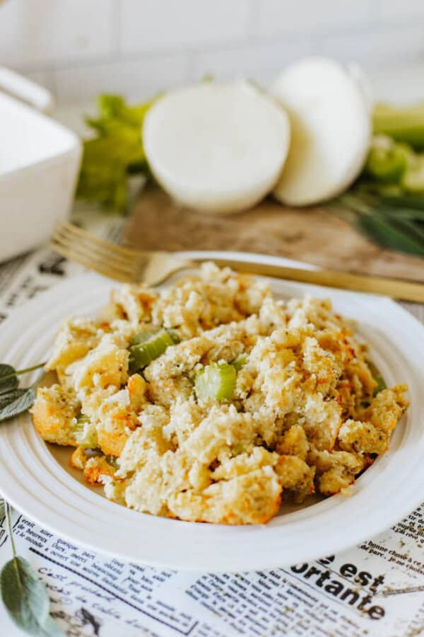 A plate of breadcrumb stuffing with celery and herbs rests on a table covered with newspaper. In the background, a halved onion and celery stalks are visible on a cutting board. A fork is placed next to the dish.
