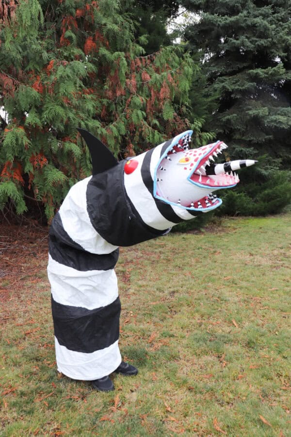 A person in a creative costume resembling a black and white striped sea creature with an open mouth and sharp teeth, standing on grass near tall evergreen trees.