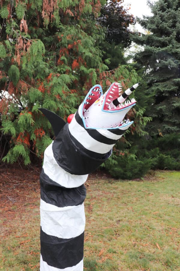 A large, striped black and white sculpture resembling a snake or worm stands upright on grassy ground. It has a jagged, open mouth with sharp red teeth and a horn, surrounded by tall trees.