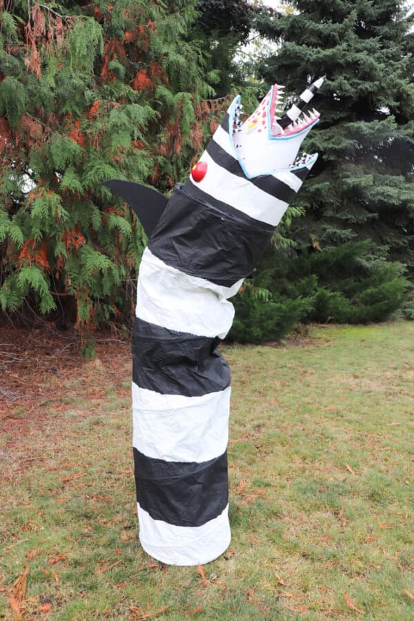 A large, striped black and white sculpture resembling a snake or worm stands upright on grassy ground. It has a jagged, open mouth with sharp red teeth and a horn, surrounded by tall trees.