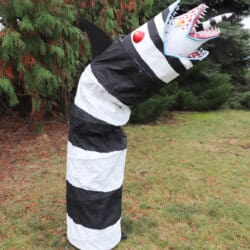 A large, striped black and white sculpture resembling a snake or worm stands upright on grassy ground. It has a jagged, open mouth with sharp red teeth and a horn, surrounded by tall trees.