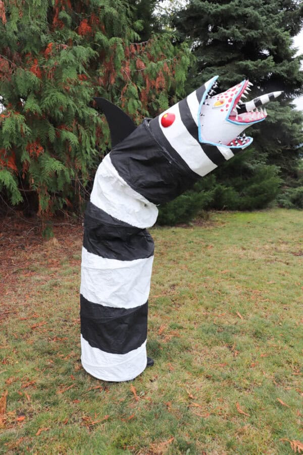 A large, striped black and white sculpture resembling a snake or worm stands upright on grassy ground. It has a jagged, open mouth with sharp red teeth and a horn, surrounded by tall trees.