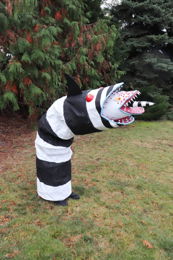 A person wearing a costume resembling a black and white striped worm with a large open mouth stands on grass, surrounded by leafy green trees. The costumes head features red lips and a pink tongue.