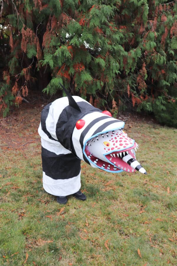 A person stands on grass wearing a costume resembling a quirky, black-and-white striped creature with red eyes and a wide mouth, featuring a protruding red tongue and spotted details. Tall, dense evergreens provide a backdrop.