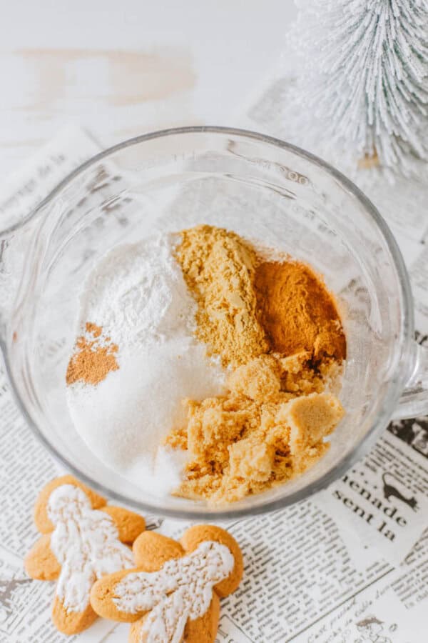 Top-down view of a glass bowl containing ingredients like sugar, flour, and spices. Two iced gingerbread cookies shaped as people lie nearby on newspaper, and a small snowy tree decorates the background.
