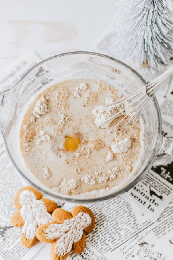 A mixing bowl with batter and an egg yolk on top, surrounded by flour, is being whisked. Two powdered gingerbread cookies are on the side, and a small, frosted artificial pine tree decorates the background.