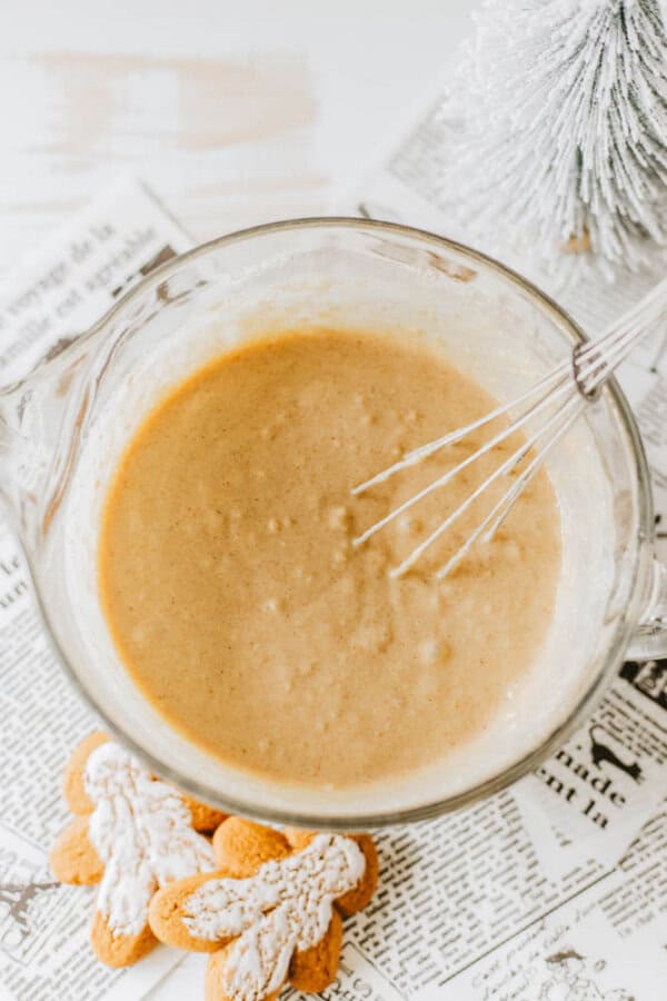 A whisk rests in a mixing bowl filled with gingerbread batter. Two gingerbread cookies with powdered sugar are placed on newspaper beneath the bowl. A small decorative, snowy tree is in the background.