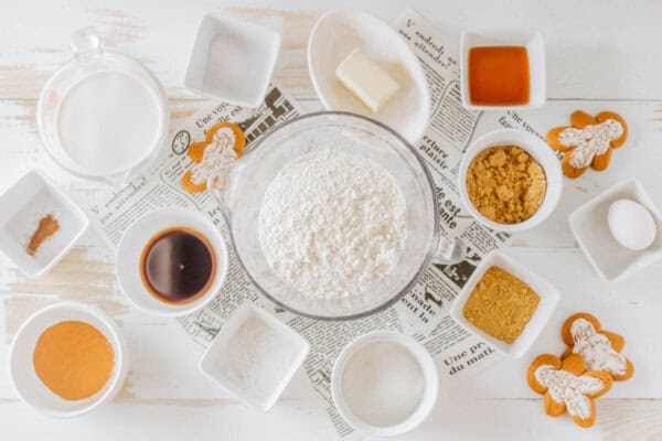 A collection of ingredients for baking gingerbread cookies arranged on a white surface. Includes flour, spices, milk, butter, sugar, honey, and eggs, with gingerbread cookies nearby. A newspaper is used as a decorative placemat.
