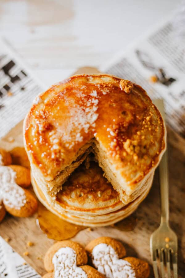 A stack of pancakes drizzled with syrup and a slice missing, resting on a wooden board. Gingerbread cookies dusted with powdered sugar surround the pancakes, and a gold fork is placed nearby. Background is blurred with newspaper pages visible.