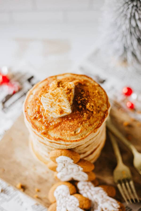 A stack of pancakes topped with a pat of butter and crumbled cookie bits sits on a wooden board. Decorative gingerbread cookies and two forks are beside the pancakes. Festive red ornaments and a small evergreen tree are in the background.