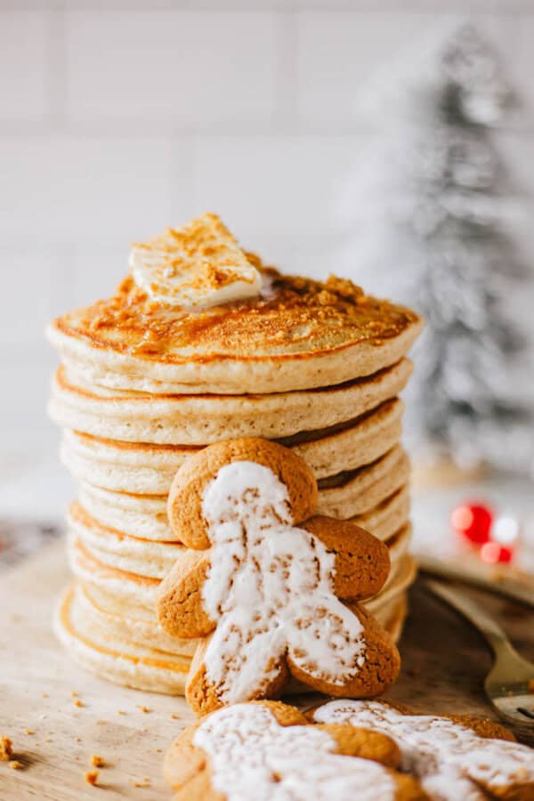 A tall stack of pancakes topped with a pat of butter, served on a wooden board. In front, there are gingerbread cookies with white icing. A blurred Christmas tree and festive decorations are in the background.
