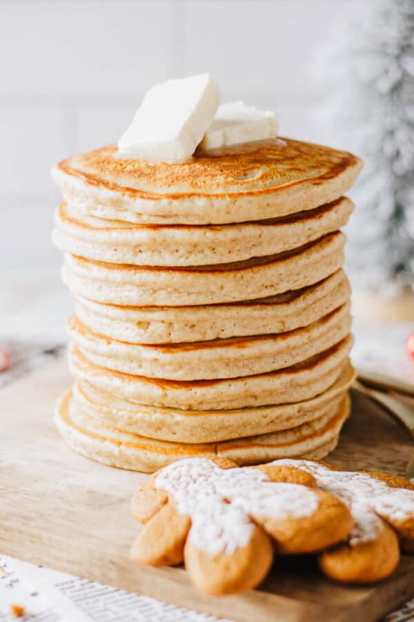 A tall stack of pancakes topped with pats of butter sits on a wooden board. In front of the pancakes are a few gingerbread cookies dusted with powdered sugar. A blurred tree is visible in the background.