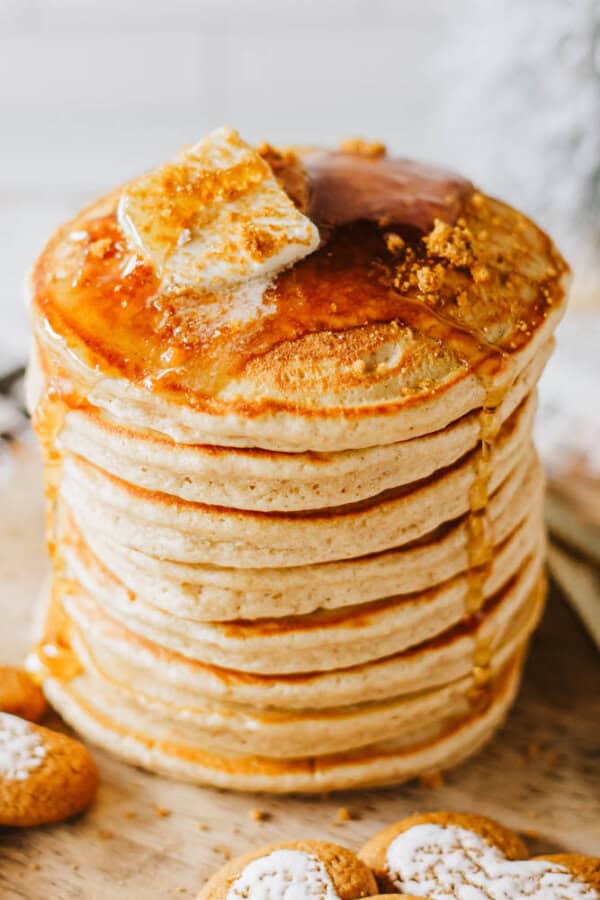 A stack of fluffy pancakes topped with melting butter and drizzled with syrup. Crumbled cookies are scattered on top and around the base, adding a decorative touch.