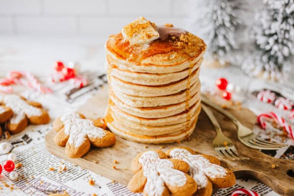 A stack of pancakes topped with syrup and a pastry square sits on a wooden board. Surrounded by gingerbread cookies, candy canes, and festive decorations, the scene evokes a cozy holiday atmosphere.
