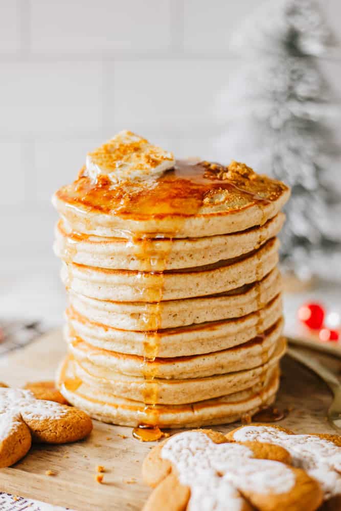 A tall stack of pancakes drizzled with syrup and topped with a slice of butter sits on a wooden board. Gingerbread cookies are in the foreground, and a blurred holiday-themed background is visible.