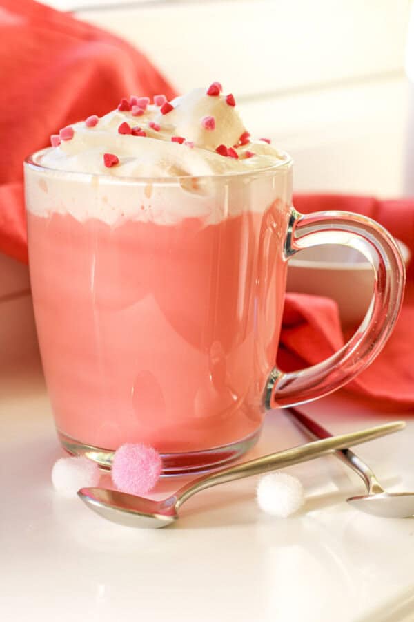 Red velvet hot chocolate in a clear mug with whipped cream and red sprinkles on top. There is a spoon and a red napkin nearby.
