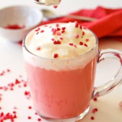 Red velvet hot chocolate in a clear mug with whipped cream and red sprinkles on top. There is a spoon with whipped cream and sprinkles, a small prep bowl with sprinkles and a red napkin nearby.
