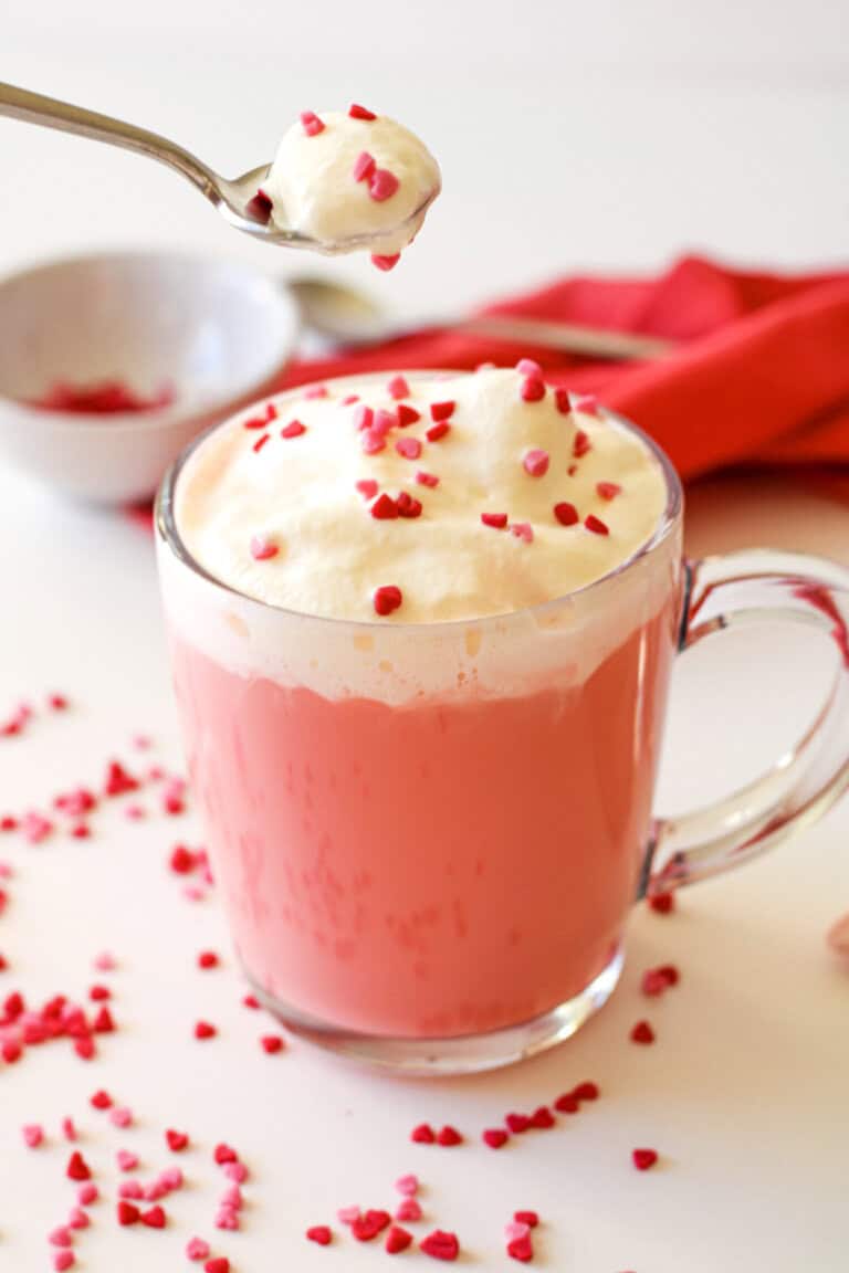 Red velvet hot chocolate in a clear mug with whipped cream and red sprinkles on top. There is a spoon with whipped cream and sprinkles, a small prep bowl with sprinkles and a red napkin nearby.