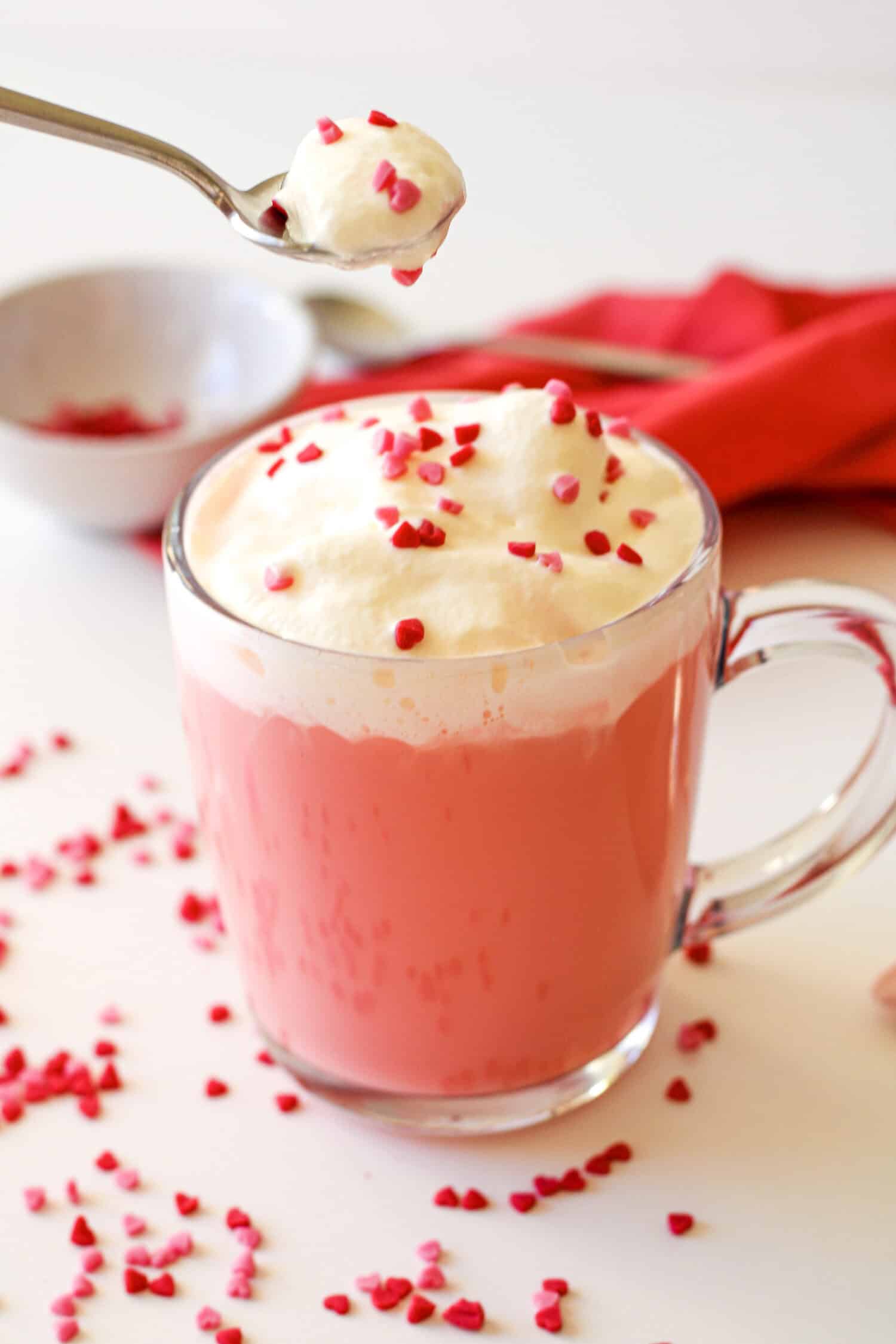 Red velvet hot chocolate in a clear mug with whipped cream and red sprinkles on top. There is a spoon with whipped cream and sprinkles, a small prep bowl with sprinkles and a red napkin nearby.