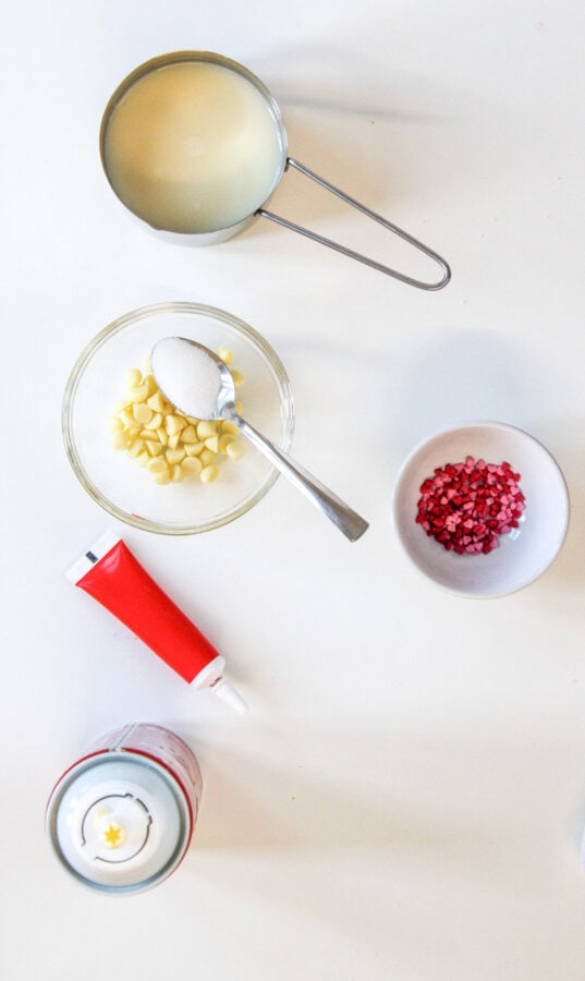 White chocolate chips in a small glass prep bowl with a spoon filled with sugar, a small prep bowl with red & pink heart shaped sprinkles, a measuring cup filled with milk, a small tube of red food colouring, and a canister of whipping cream.