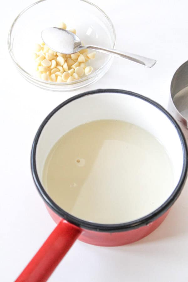Milk in a red pot, white chocolate chips in a small prpe bowl with a spoon in it.