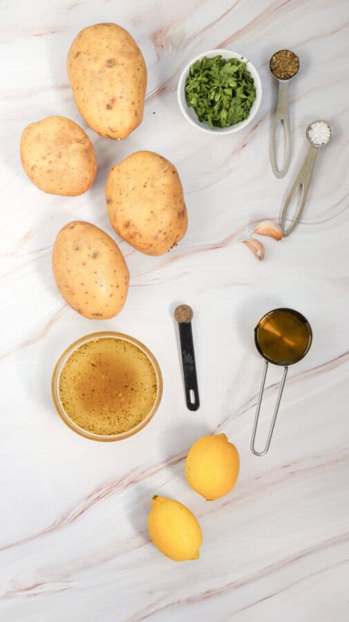 Raw whole potatoes, whole lemons, garlic cloves, parsley in a prep bowl, vegetable broth in a glass prep bowl, salt & pepper in measuring spoons, oil in a measuring cup.