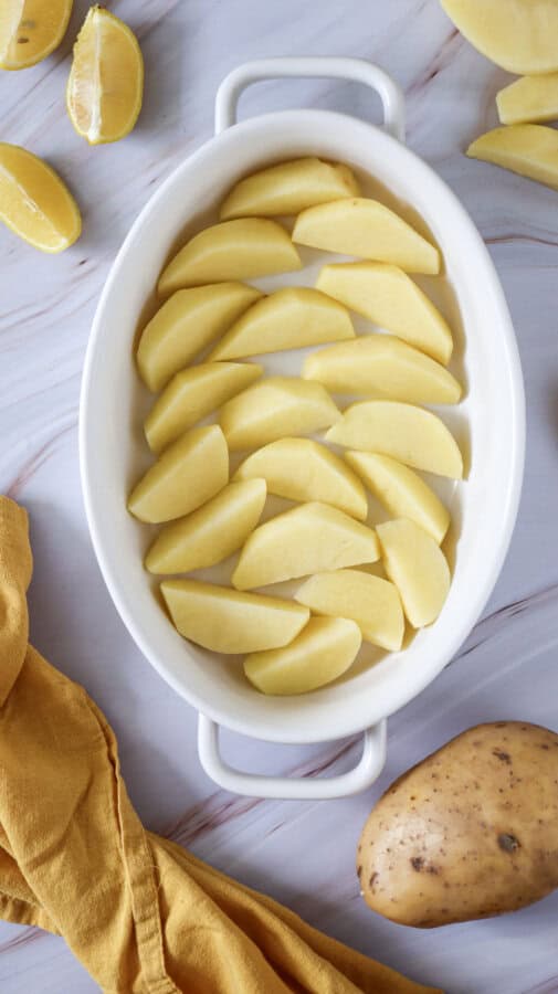 Raw, cut up potatoes in a white baking dish surrounded by another whole potatoes, some lemon wedges and a yellow napkin.