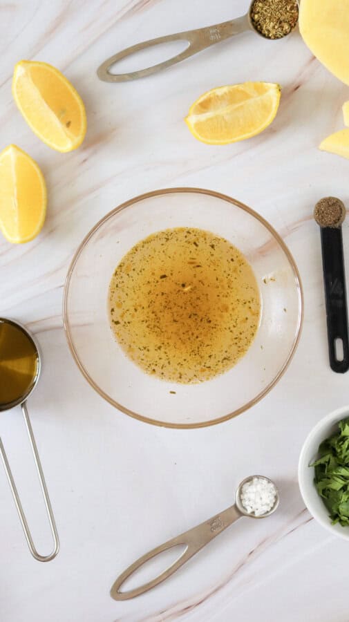 Lemon juice, olive oil and vegetable broth mixed in a small glass bowl, with lemon wedges and measuring spoons with spices surrounding.