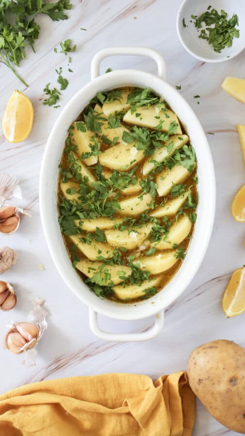 Sliced potatoes in covered in sauce with parsley on top in a white baking dish, with garlic cloves and lemon wedges surrounding.