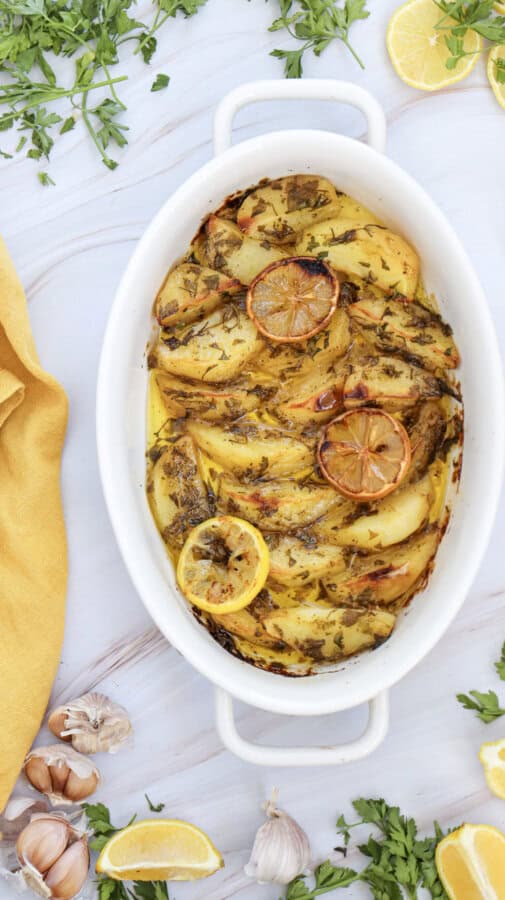 Lemon greek potatoes in a white casserole dish garnished with fresh parsley and with herbs, garlic cloves and lemons and a yellow napkin surrounding it.