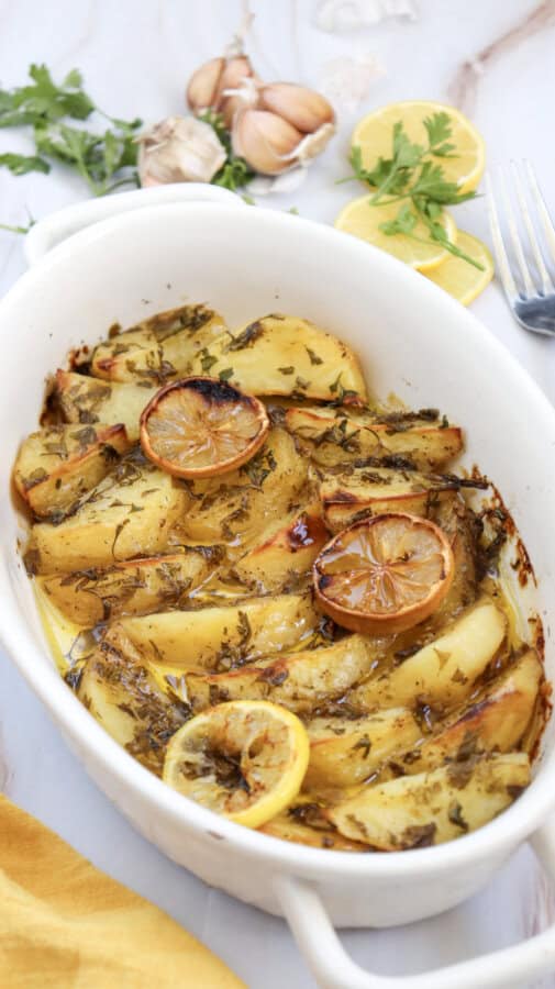 Lemon greek potatoes garnished with parsley and lemon slices on top in a white baking dish, with garlic cloves, sliced lemons and sprigs of parsley and a fork surrounding.