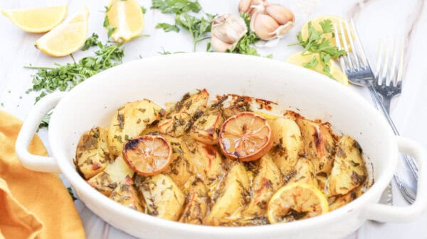 Lemon greek potatoes in a white casserole dish. surrounded by fresh parsley, sliced lemons, garlic cloves, an orange napkin, and two forks.