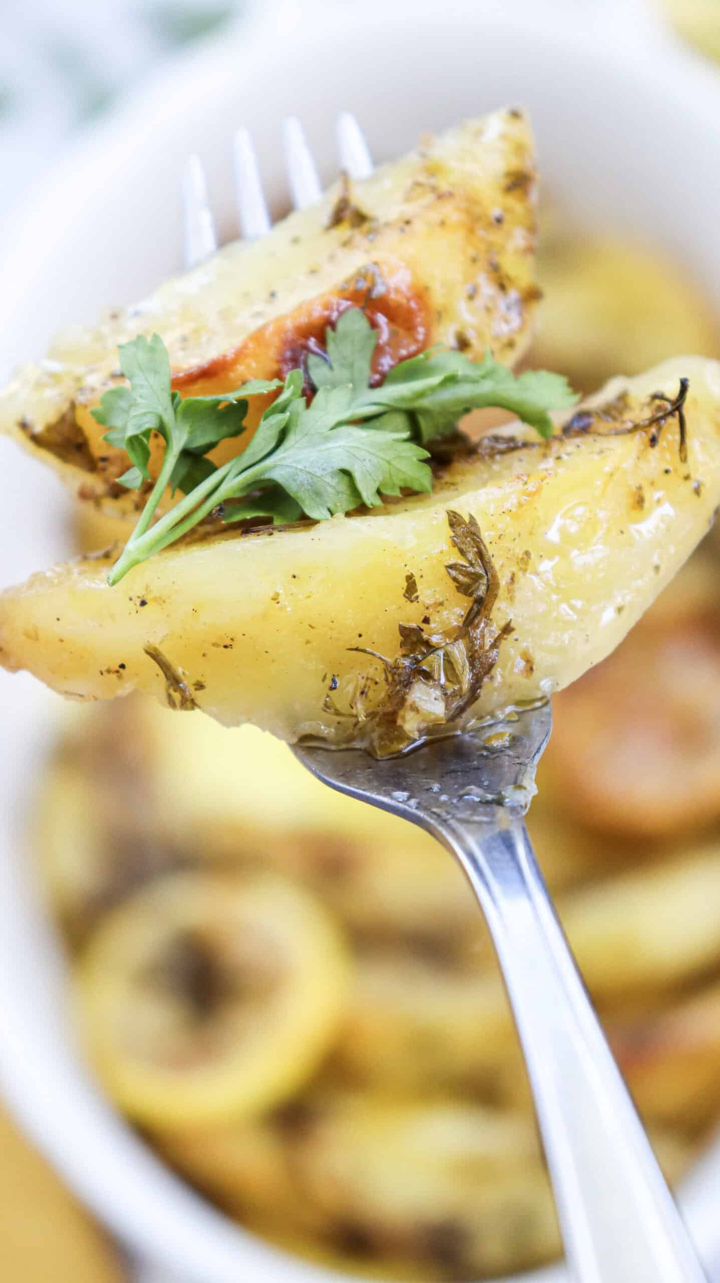 Lemon greek potatoes on a fork, garnished with fresh parsley, with a casserole dish of potatoes in the background.