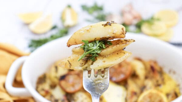 Lemon greek potatoes on a fork, garnished with fresh parsley, with a casserole dish of potatoes in the background.