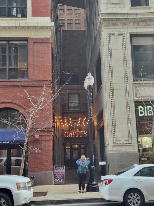 A person stands on the sidewalk looking at a narrow alleyway between two tall buildings. At the end of the alley, a lit sign reads Coffee with bright marquee letters. The aroma of homemade treats drifts through the air, as white cars are parked along the street.