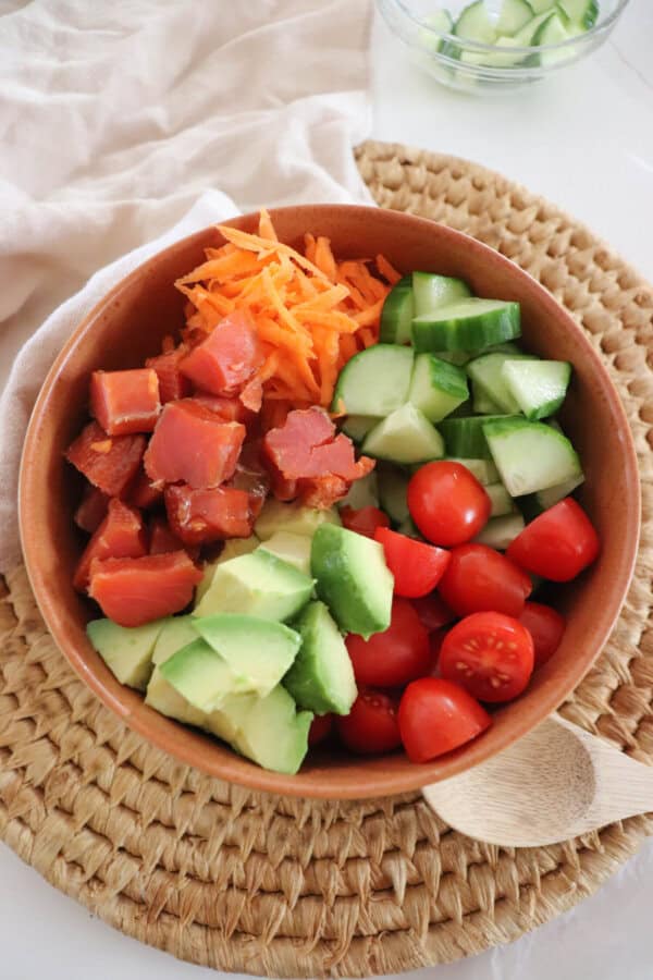 A colorful rice bowl featuring diced smoked salmon, sliced cucumbers, halved cherry tomatoes, shredded carrots, avocado chunks, all in a wooden bowl on a woven mat.