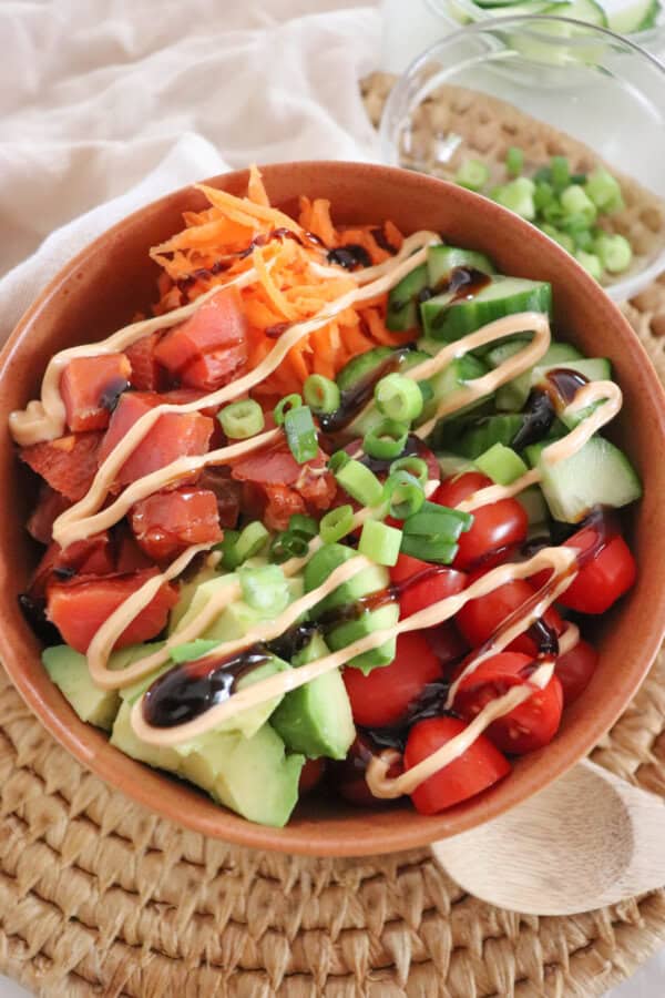 A colorful rice bowl featuring diced smoked salmon, sliced cucumbers, halved cherry tomatoes, shredded carrots, avocado chunks, and a drizzle of spicy mayonnaise an teriyaki sauce garnished with chopped green onions and sesame seeds, all in a wooden bowl on a woven mat.