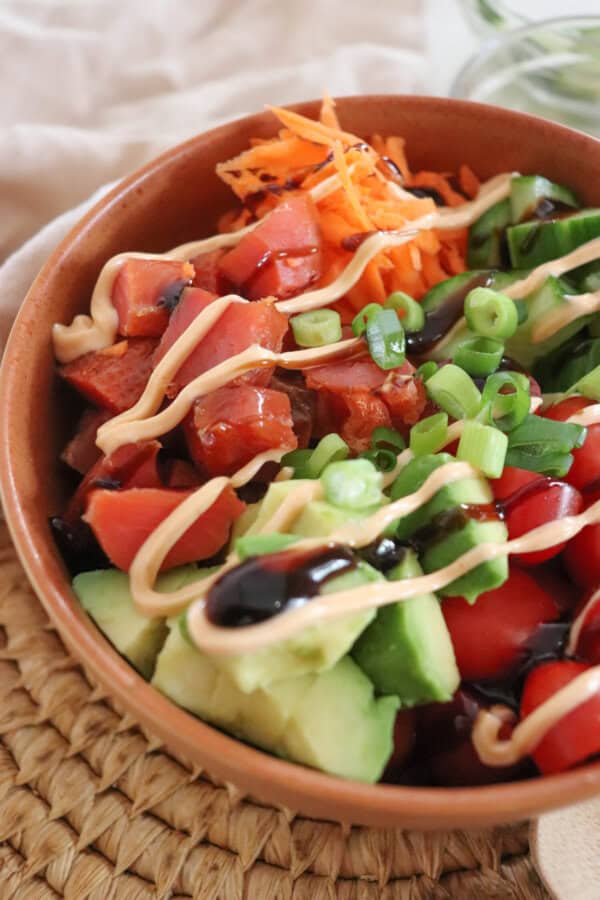 A colorful rice bowl featuring diced smoked salmon, sliced cucumbers, halved cherry tomatoes, shredded carrots, avocado chunks, and a drizzle of spicy mayonnaise an teriyaki sauce garnished with chopped green onions, all in a wooden bowl on a woven mat.