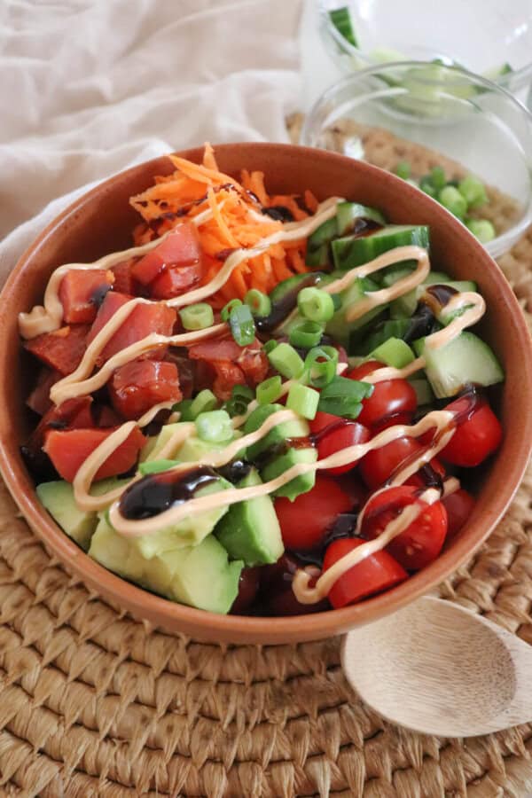 A colorful rice bowl featuring diced smoked salmon, sliced cucumbers, halved cherry tomatoes, shredded carrots, avocado chunks, and a drizzle of spicy mayonnaise an teriyaki sauce garnished with chopped green onions, all in a wooden bowl on a woven mat with a wooden spoon and some green onions in a clear prep bowl on the side.