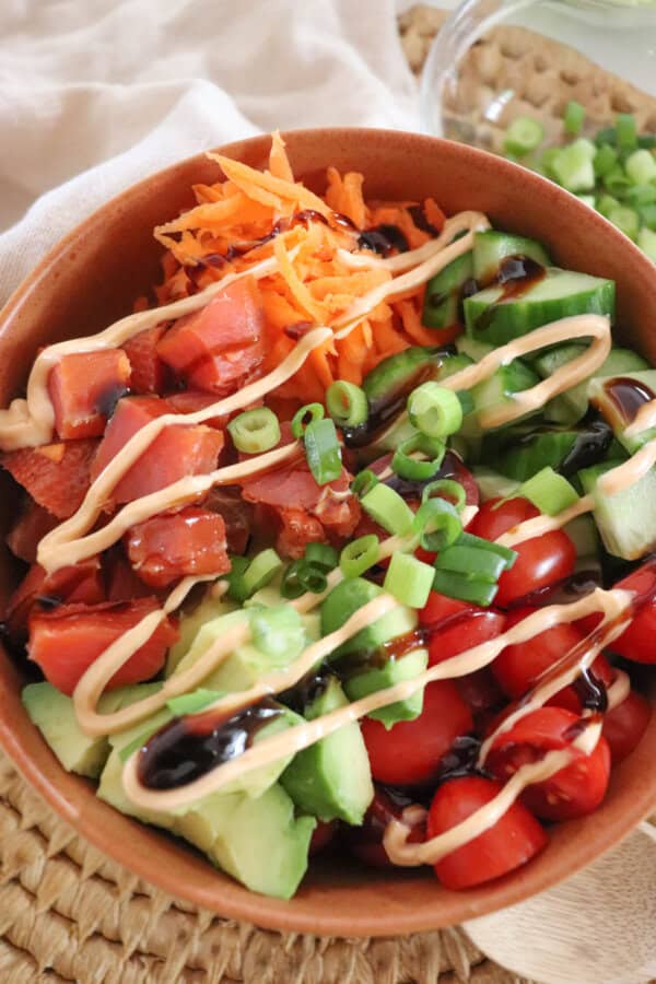 A colorful rice bowl featuring diced smoked salmon, sliced cucumbers, halved cherry tomatoes, shredded carrots, avocado chunks, and a drizzle of spicy mayonnaise an teriyaki sauce garnished with chopped green onions, all in a wooden bowl on a woven mat.