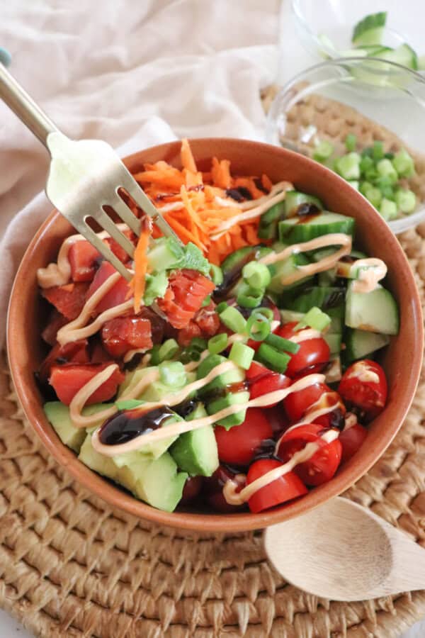 A colorful rice bowl featuring diced smoked salmon, sliced cucumbers, halved cherry tomatoes, shredded carrots, avocado chunks, and a drizzle of spicy mayonnaise an teriyaki sauce garnished with chopped green onions, all in a wooden bowl on a woven mat with a fork.