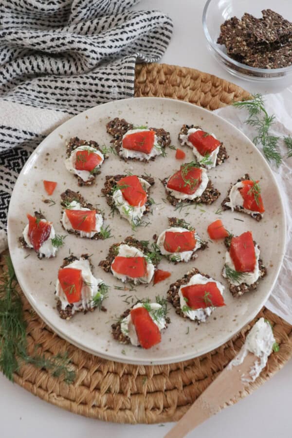 Crackers with cream cheese and pieces of smoked salmon garnished with fresh dill on a cream plate on a woven mat, with a small prep bowl with more crackers, a sprig of dill and a tea towel surrounding.