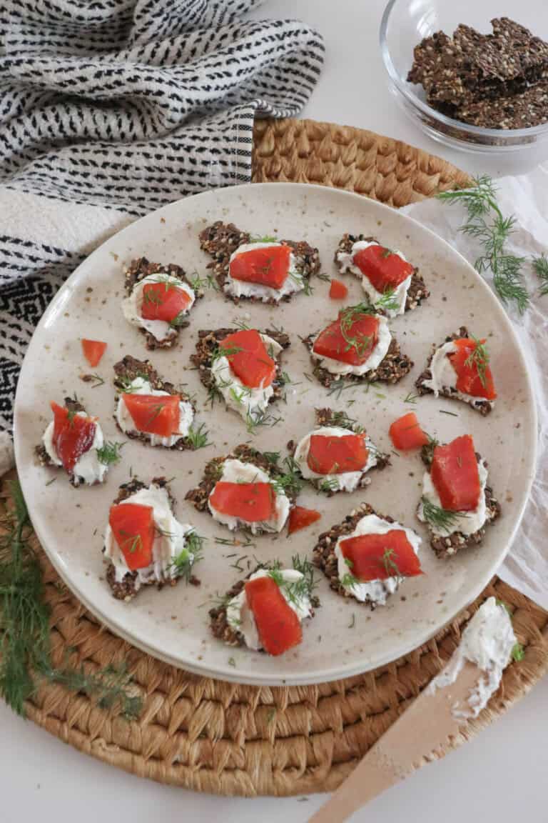 Crackers with cream cheese and pieces of smoked salmon garnished with fresh dill on a cream plate on a woven mat, with a small prep bowl with more crackers, a sprig of dill and a tea towel surrounding.