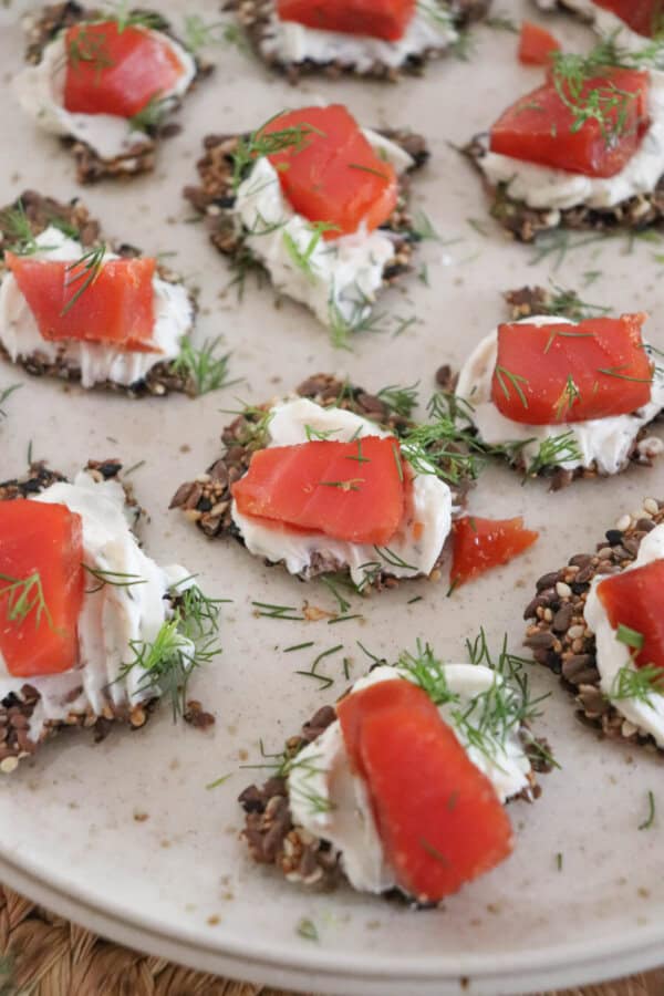 Bites of salmon on cream cheese and crackers on a cream plate with dill garnish.