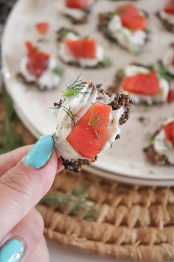 Someone holding a cracker with cream cheese and pieces of smoked salmon garnished with fresh dill with more crackers on a cream plate and a woven mat.