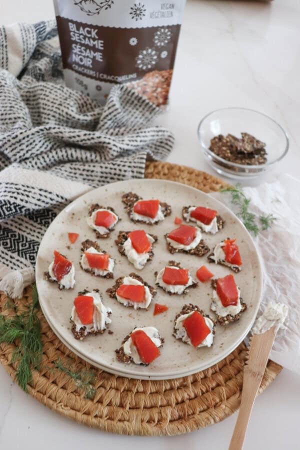 Crackers with cream cheese and pieces of smoked salmon garnished with fresh dill on a cream plate on a woven mat, with a small prep bowl with more crackers, a sprig of dill, a bag of Eve's Crackers and a tea towel surrounding.