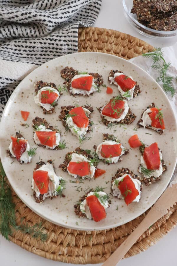 Crackers with cream cheese and pieces of smoked salmon garnished with fresh dill on a cream plate on a woven mat, with a small prep bowl with more crackers, a sprig of dill and a tea towel surrounding.